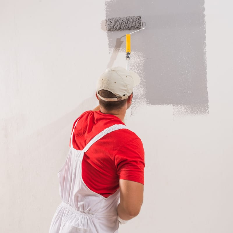 Interior painting in progress â€” a professional wearing white overalls and a cap uses a paint roller to apply gray paint to a wall, representing expert wall finishing as part of a residential renovation.
